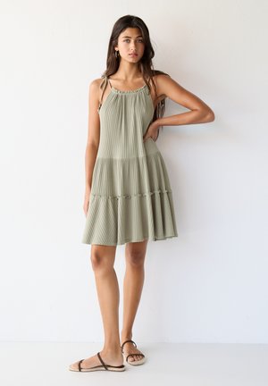 Abito plissettato verde chiaro con orlo arricciato, spalline regolabili e silhouette morbida. Abbinato a semplici sandali marroni.