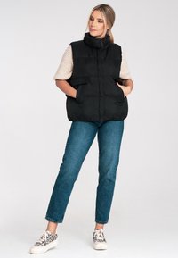 Czarna puchowa kamizelka z wysokim kołnierzem, dużymi przednimi kieszeniami i pikowanym wzorem, w połączeniu z jasnym swetrem i niebieskimi dżinsami.