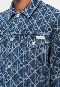 Jeans jakna s geometrijskim plavim uzorkom, s aluminijskim gumbima i istaknutim Calvin Klein logom na prsima.