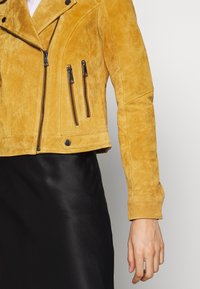 Veste en daim jaune avec plusieurs fermetures éclair verticales portée sur une jupe noire, montrant un gros plan du bras droit et du torse.