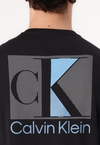 Black T-shirt featuring a graphic design on the back with perforated pattern. Includes large white "K" and light blue "Calvin Klein" text.
