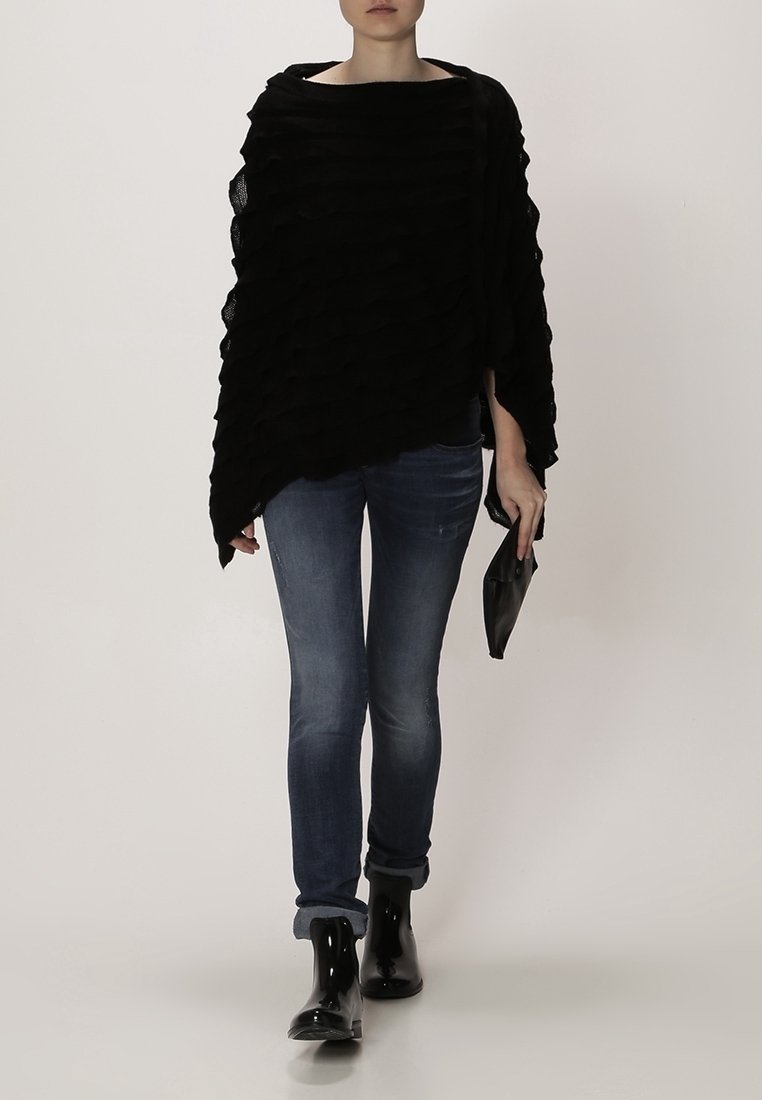Svart texturerad poncho med vågig design, bärs över åtsittande jeans, kompletterad med glansiga svarta ankelstövlar och en clutchväska.