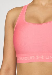 Rózsaszín sportmelltartó sima anyagból, mély kivágással, hátul versenyző fazonú pánttal és az elasztikus szalagon található Under Armour logóval.