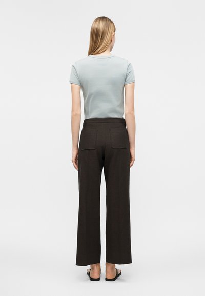 Γυναίκα που στέκεται με την πλάτη γυρισμένη φοράει ανοιχτό μπλε μπλουζάκι με κοντά μανίκια, μαύρο παντελόνι φαρδύ με τσέπες στην πλάτη και μαύρα σανδάλια σε λευκό φόντο.