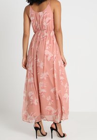 Maxiklänning i mjuk rosa tyg med vita blommönster, som har en insvängd midja, justerbara spaghettiband och en svävande silhuett.