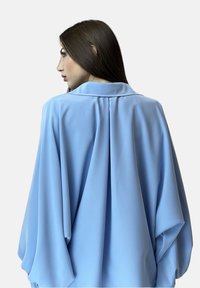 Blusa azul clara con una tela suave y fluida. Presenta un cuello clásico, mangas drapeadas y un detalle de plisado en la parte posterior.