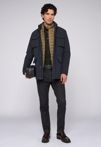 Granatowa kurtka wierzchnia z wieloma kieszeniami na beżowym ocieplanym płaszczu, zielona koszula z guzikami, ciemnoniebieskie dżinsy i brązowe buty. Czarna torba trzymana w ręce.