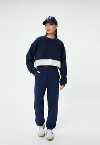 Sudadera recortada azul marino con dobladillo blanco, combinada con joggers azul marino a juego. Los accesorios incluyen una gorra azul marino y zapatillas blancas con detalles en negro.