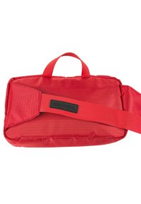 Bolsa de tela roja con forma rectangular plana, que cuenta con un asa, correa ajustable y un parche de logo negro. Superficie texturizada.