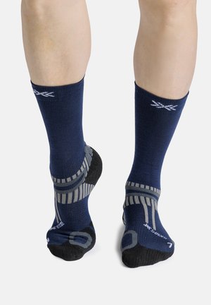 Benen met marineblauwe atletische crew-sokken met grijze en zwarte accenten, gezien van achteren tegen een witte achtergrond.