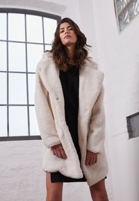 Cappotto in finta pelliccia di colore crema con cappuccio, caratterizzato da una superficie morbida e testurizzata, vestibilità ampia e chiusura con un solo bottone. Indossato sopra un vestito scuro.