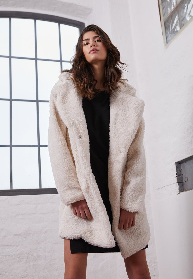 Cappotto in finta pelliccia di colore crema con cappuccio, caratterizzato da una superficie morbida e testurizzata, vestibilità ampia e chiusura con un solo bottone. Indossato sopra un vestito scuro.