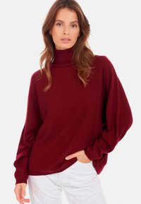 Pull en col roulé bordeaux avec des manches amples, fabriqué en maille douce, associé à un pantalon blanc, offrant une silhouette décontractée.