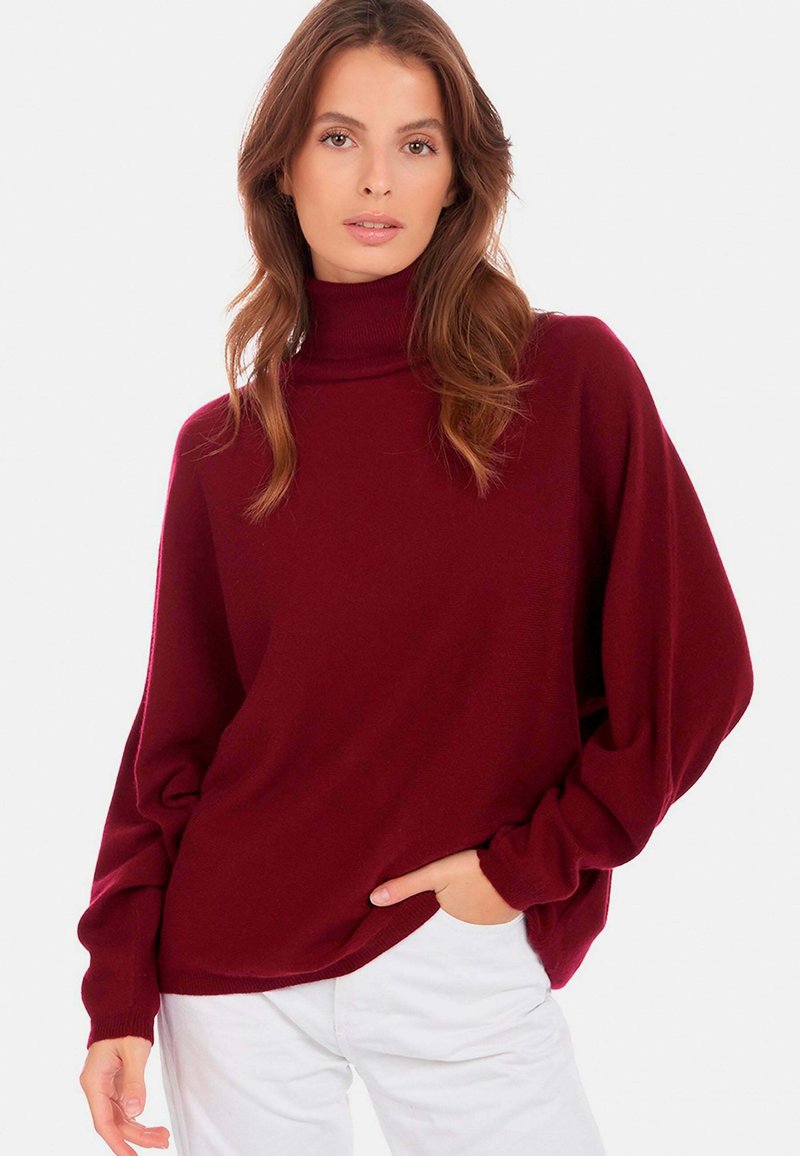 Pull en col roulé bordeaux avec des manches amples, fabriqué en maille douce, associé à un pantalon blanc, offrant une silhouette décontractée.