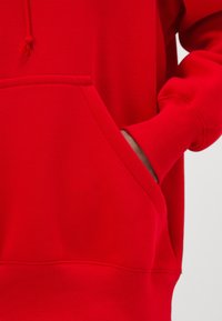 Personne portant un sweat à capuche rouge vif avec une main partiellement dans la poche kangourou avant, sur un fond clair.