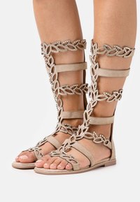 Beige gladiator sandals yang terbuat dari bahan suede, memiliki pola rantai dekoratif dengan aksen rhinestone, dan sol datar.