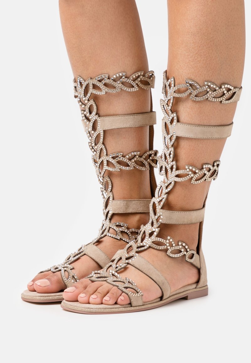 Beige gladiator sandals yang terbuat dari bahan suede, memiliki pola rantai dekoratif dengan aksen rhinestone, dan sol datar.