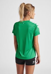 Grøn kortærmet sports skjorte med en glat tekstur, prydet med hvide chevron-detaljer på skuldrene og en rund halsudskæring.