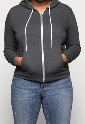 Personne portant un sweat à capuche zippé gris foncé avec des cordons blancs et un jean bleu, les mains dans les poches avant, debout devant un fond uni.