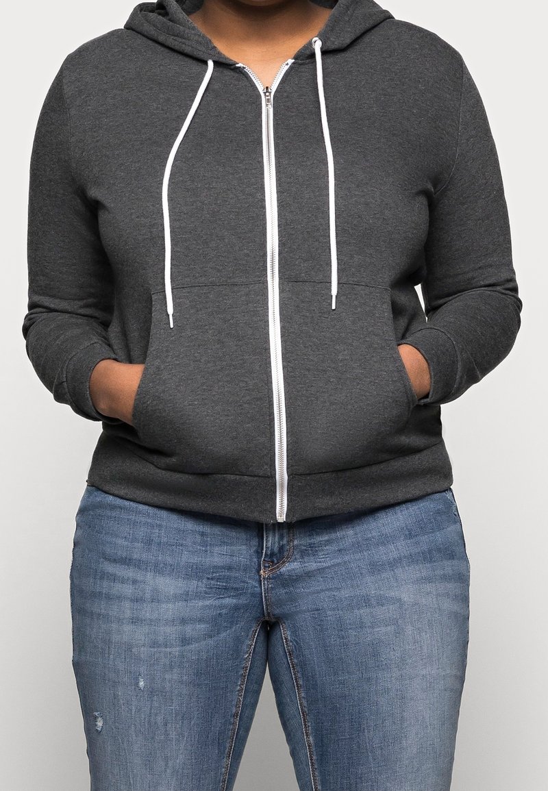 Personne portant un sweat à capuche zippé gris foncé avec des cordons blancs et un jean bleu, les mains dans les poches avant, debout devant un fond uni.