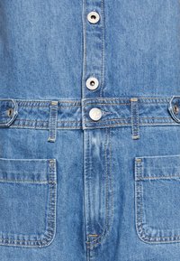 Denimski kombinezon s svetlo modrim pranjem, s srebrnimi gumbi na pritisak, šivalnimi detajli in dvema sprednjima žepoma na blazini in straneh.