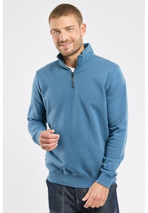 Homme souriant portant un pull à demi-fermeture éclair bleu et un jean foncé, debout devant un fond blanc uni.