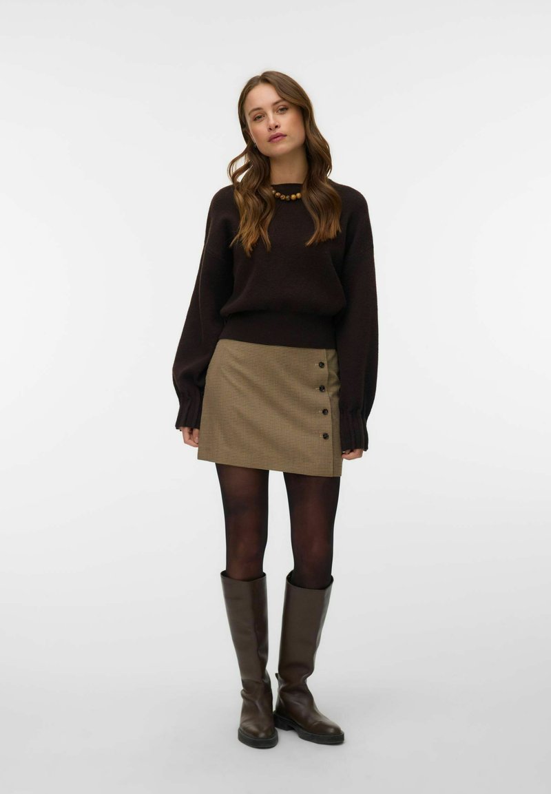 Femme aux cheveux ondulés portant un pull marron foncé, une mini-jupe beige boutonnée, des collants foncés et de grandes bottes marron, debout sur un fond blanc.