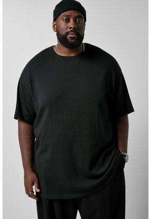 Man draagt een zwarte beanie, zwart oversized T-shirt, kettingketting, ring en polshorloge, staande tegen een effen lichte achtergrond.