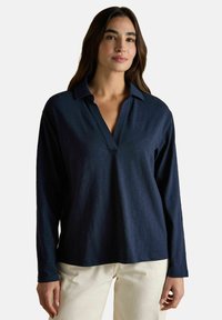 Naine kannab tumehallide pikade varrukatega polo-särki avatud kaelusega ja beeže pükse, seistes ühtlase tausta ees.