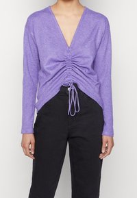 Lawendowy sweter z głębokim dekoltem w serek i marszczonym detalem na przodzie. Długie rękawy i regulowany ściągacz u dołu. Tkanina dzianinowa o fakturze.