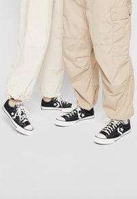 Baskets en toile noire avec semelles en caoutchouc blanches, arborant un motif d'étoile blanche, associées à un pantalon cargo beige et un pantalon en tissu léger.