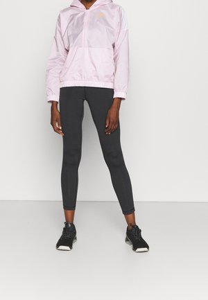 Persona con chaqueta con capucha rosa claro, leggings ajustados negros y zapatos deportivos negros de pie frente a un fondo blanco liso.