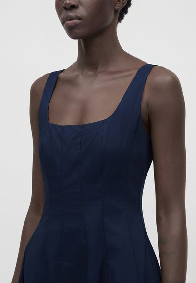 Femme à la peau foncée portant une robe bleu marine cintrée sans manches avec un col carré sur un fond clair uni.