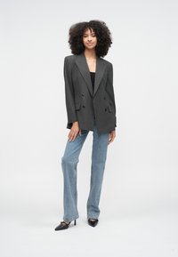 Žena s kučeravými vlasmi, oblečená v tmavom oversized blejzri s tenkými pásikmi, čiernom topu, svetlomodrých džínsoch a čiernych špicatých lodičkách, stojaca pred bielym pozadím.