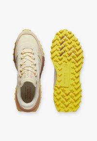 Las zapatillas Lacoste presentan un upper beige con acentos en rosa, una suela de goma amarilla texturizada con un patrón de agarre multidireccional.