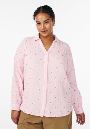 Žena kučeravých vlasov, oblečená v svetloružovej blúzke na gombíky s malými vyšívanými kvetmi a béžových nohaviciach, stojaca na bielom pozadí.