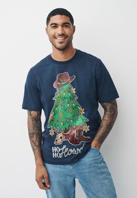 Tamsiai mėlynas marškinėlis su spalvinga dekoruoto Kalėdų medžio, kaubojaus skrybėlės ir aulinukų grafika, o po ja užrašas „Ho Ho Howdy“.