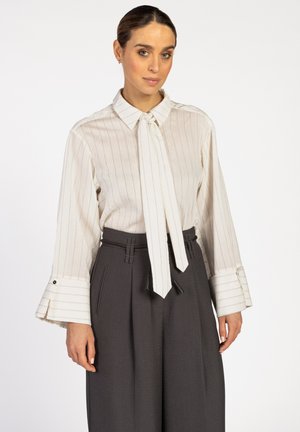 Vrouw in een witte blouse met lange mouwen met verticale strepen en brede manchetten, gecombineerd met hoge, donkergrijze, geplooide broeken.