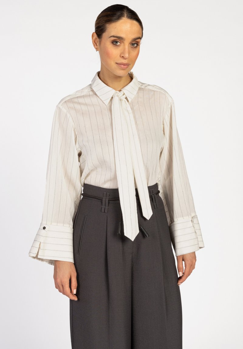 Vrouw in een witte blouse met lange mouwen met verticale strepen en brede manchetten, gecombineerd met hoge, donkergrijze, geplooide broeken.