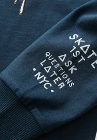Sötétkék pulóver, texturált felülettel. Fehér nyomtatott szöveggel rendelkezik: "ELŐBB DESZKÁZZ, AZTÁN KÉRDEZZ" és "NYC" geometrikus díszítésekkel.