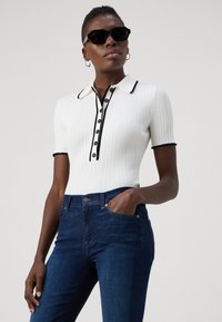 Witte geribde polo met zwarte rand en knopen, gecombineerd met donkerblauwe denim jeans. Model draagt zwarte zonnebril en zilveren hoepel oorbellen.