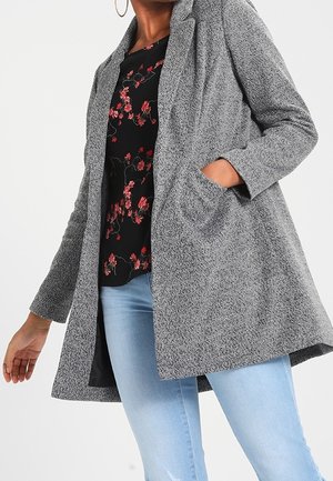 Vrouw draagt een grijze open jas over een zwarte top met bloemenprint en lichtblauwe spijkerbroek, met één hand in de jaszak en zichtbare hoepeloorringen.