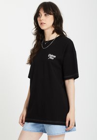 Czarny bawełniany t-shirt o luźnym kroju, z krótkimi rękawami, z białym nadrukiem logo i kontrastowym przeszyciem. W połączeniu z jasnoniebieskimi szortami dżinsowymi.