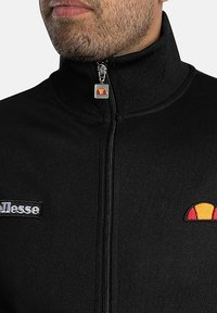 Chaqueta de forro polar negra con cremallera y cuello alto. Presenta un logotipo bordado y un acento colorido cerca de la cremallera. Tiene una textura suave y un ajuste ceñido.