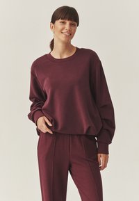 Bordeaux sweatshirt met een ronde halslijn en lange mouwen, gecombineerd met bijpassende broek met voorste plooidetails. Zachte stoftextuur.