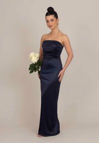 Pánt nélküli tengerészkék szatén ruhaköltemény, testhez simuló fazonnal, a felsőrészen ráncolt részlettel és sima textúrával. A modell fehér rózsákat tart a kezében.