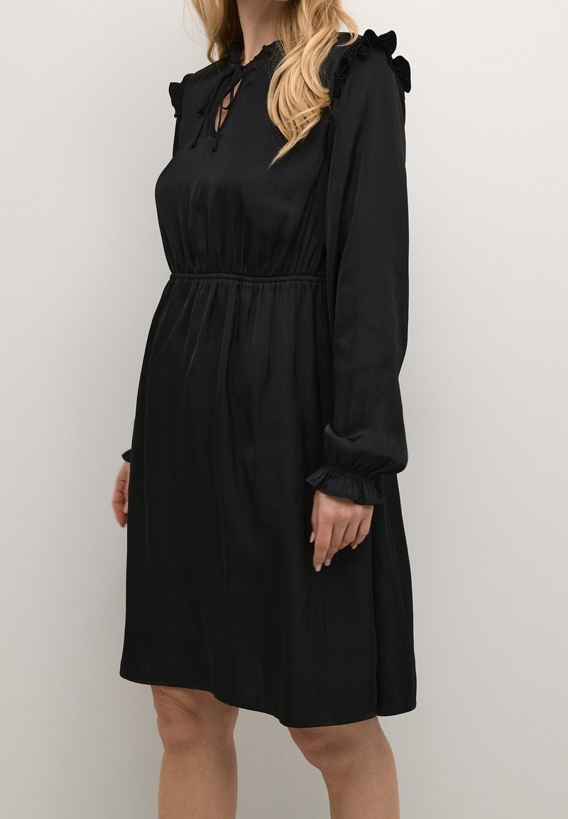 Svart klänning med hög krage, volangdetaljer på axlarna, långa ärmar och inringad midja. Tillverkad av ett mjukt tyg, knälång.