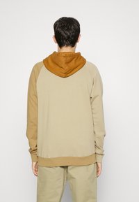 Sweat à capuche avec un corps beige et une capuche marron. Confectionné en tissu doux, avec des poignets et un ourlet côtelés pour une finition ajustée.