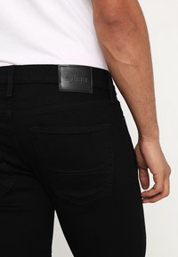 Svarta jeans har en slank, skräddarsydd design med bakfickor och ett läderlogomärke på midjebandet. Tyget har en mjuk textur.