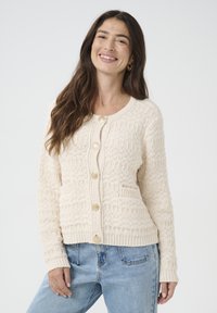 Cardigan bej tricotat cu un model texturat, guler rotund, buzunare frontale și nasturi aurii. Asortat cu blugi albaștri deschiși.